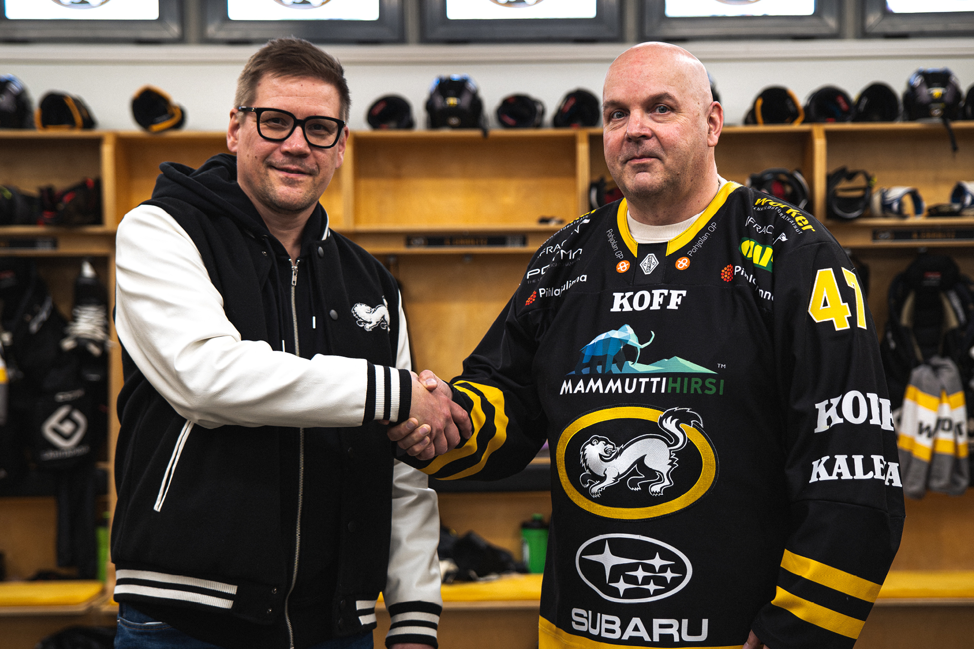 Kaksi miestä katsoo kameraan ja kättelee. Toisella miehellä on Kärpät-pelipaita päällä ja he ovat jäähallin pukuhuoneessa.
